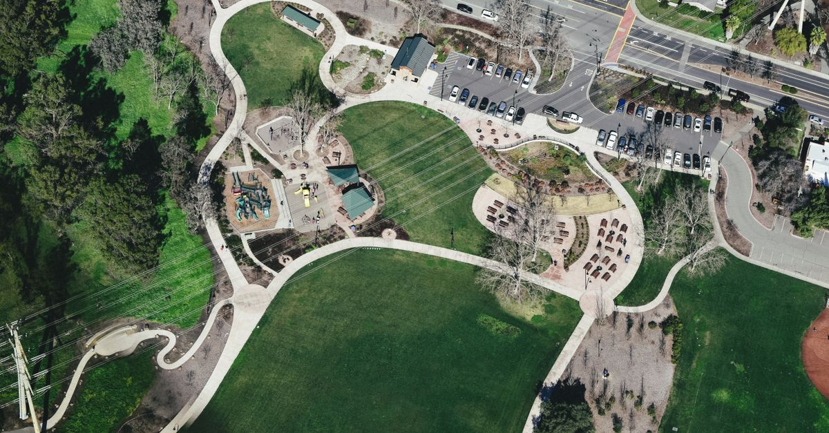 Drone view of modern city park area with winding roads surrounded by greenery and cars parked nearby located under power lines